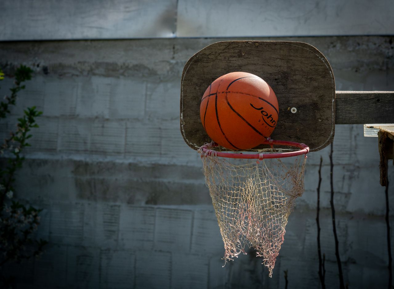 about-us Close-up of a basketball on an outdoor hoop with a worn net and wooden backboard.